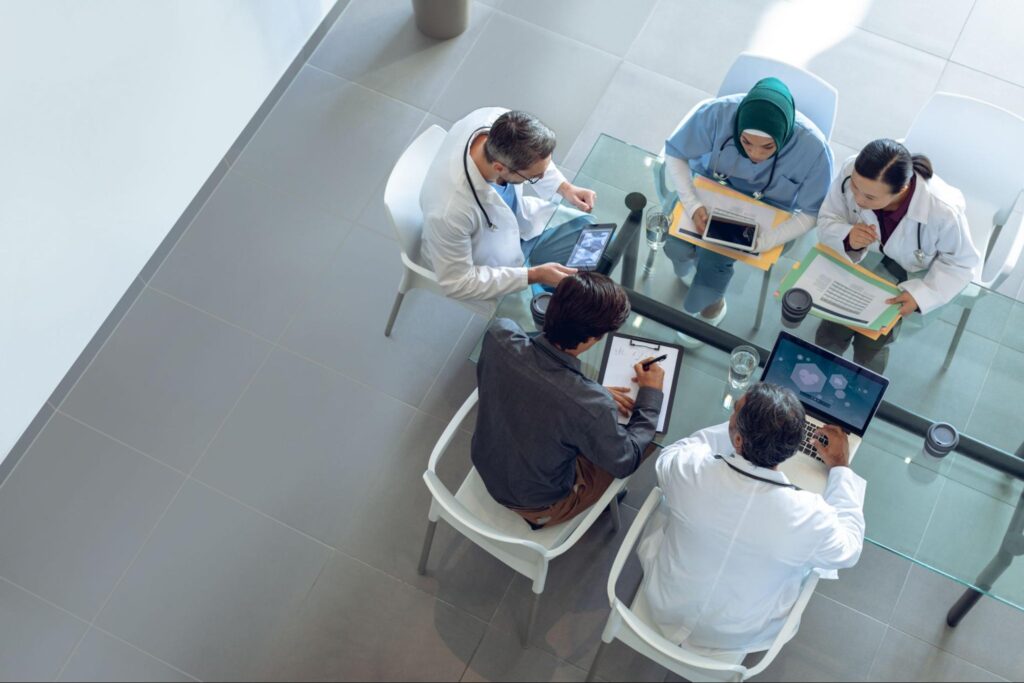 An aerial view of a group of five physicians sitting at a glass conference table.