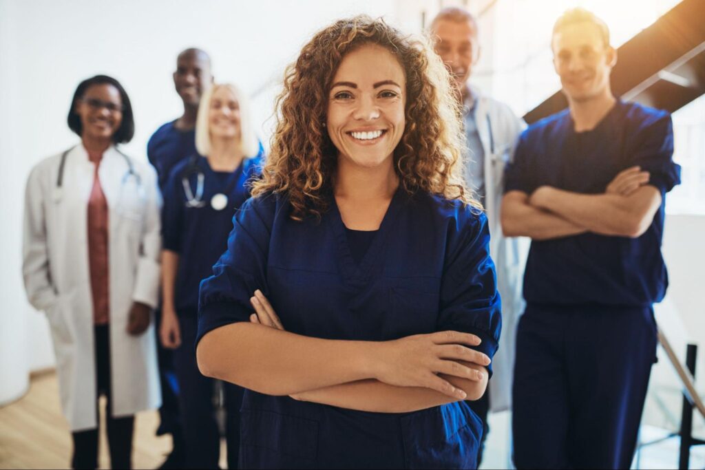 A group of physicians stand in front of a staircase. A young woman with curly hair stands, smiling, at the front, while the five other individuals are slightly blurred in the background.